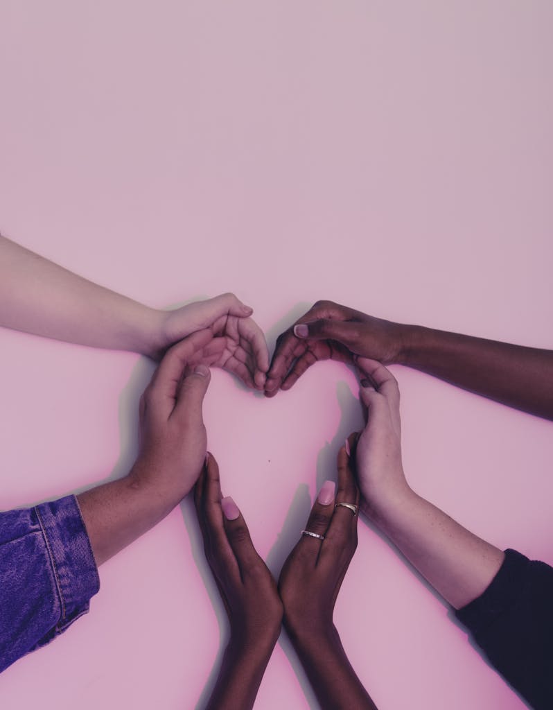 A group of diverse hands forming a heart shape against a soft pink background, symbolizing love and unity.