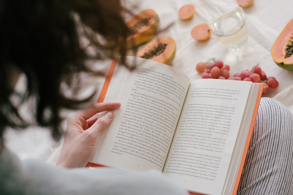 A person enjoys reading a book surrounded by fresh papaya, grapes, and a drink, creating a cozy indoor scene.